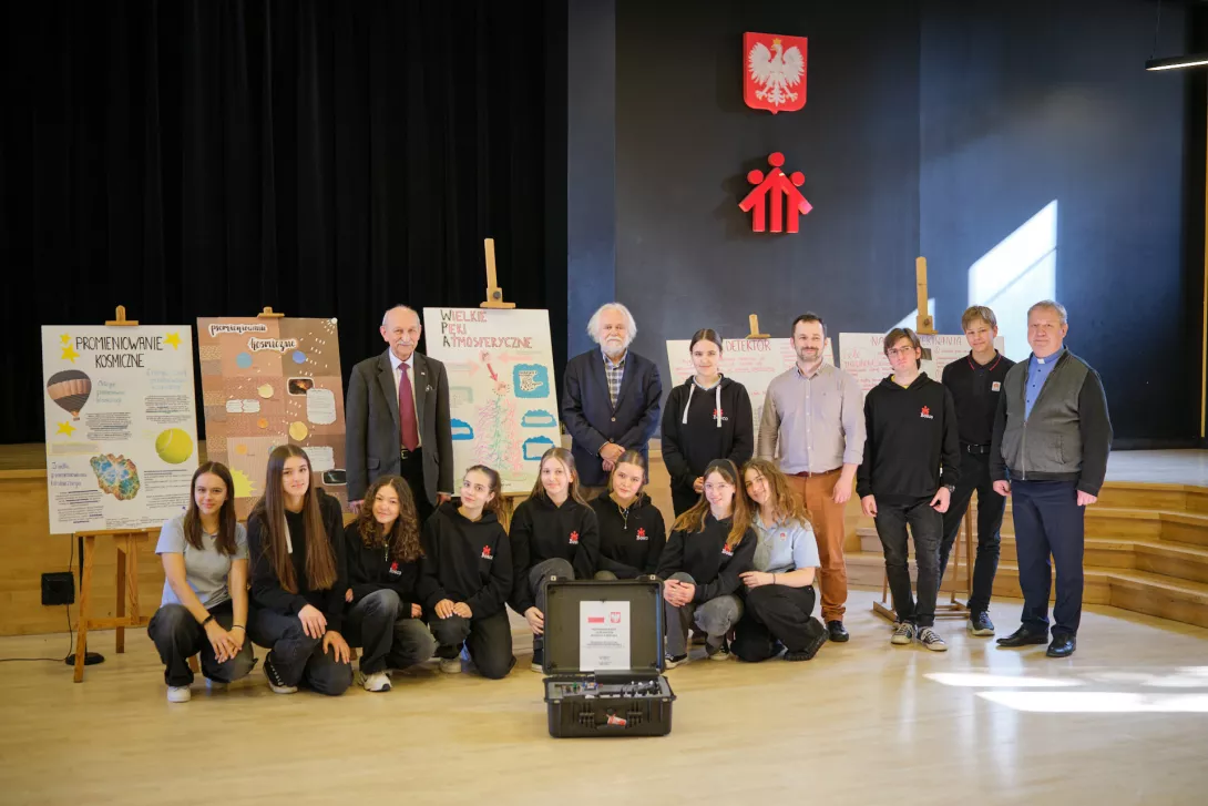 Cała grupa zaangażowana w projekt: uczniowie, p. Czekalski, prof. Wibig, dyrektor na tle plakatów o promieniowaniu kosmicznym. (fot. Amelia Szymańska)