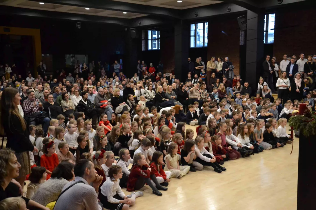 Rodzice i dzieci zgromadzeni w auli szkolnej w czasie świątecznego spotkania dla klas wczesnoszkolnych. 