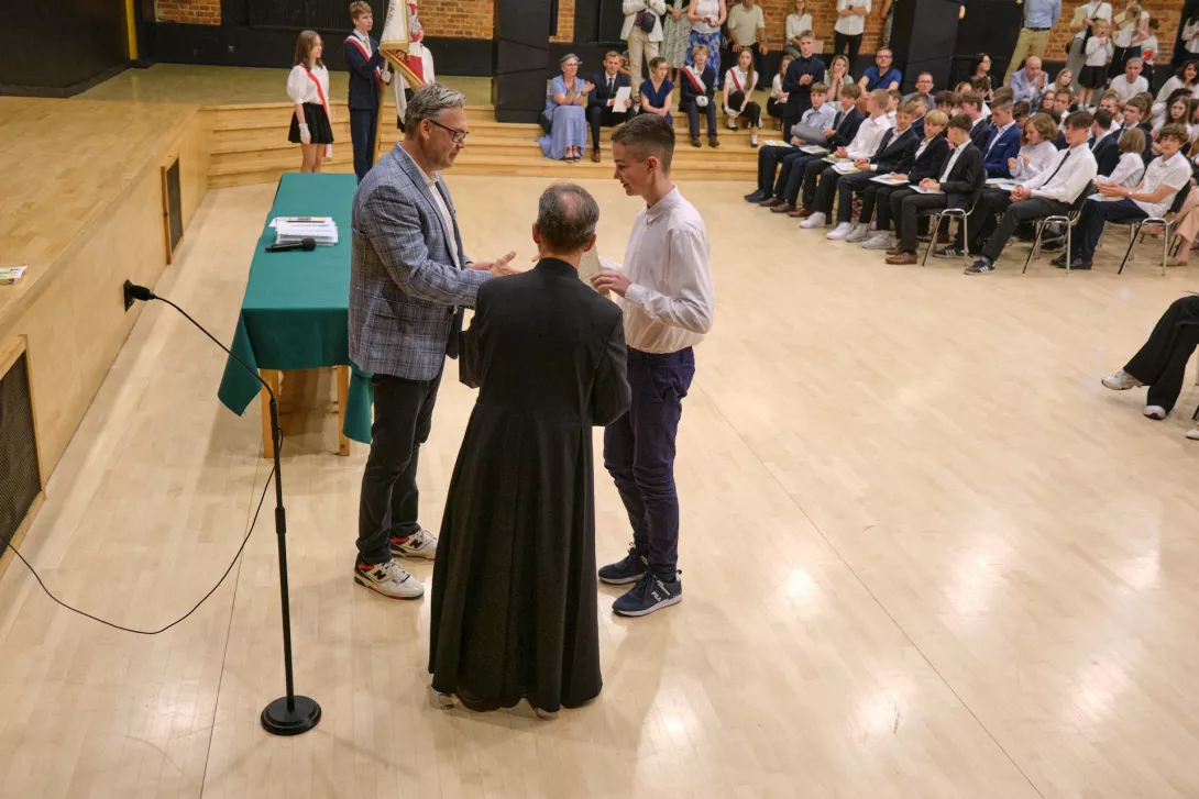 Wojtek odbiera nagrodę "Godny naśladowania" od wychowawcy i ks. dyrektora.